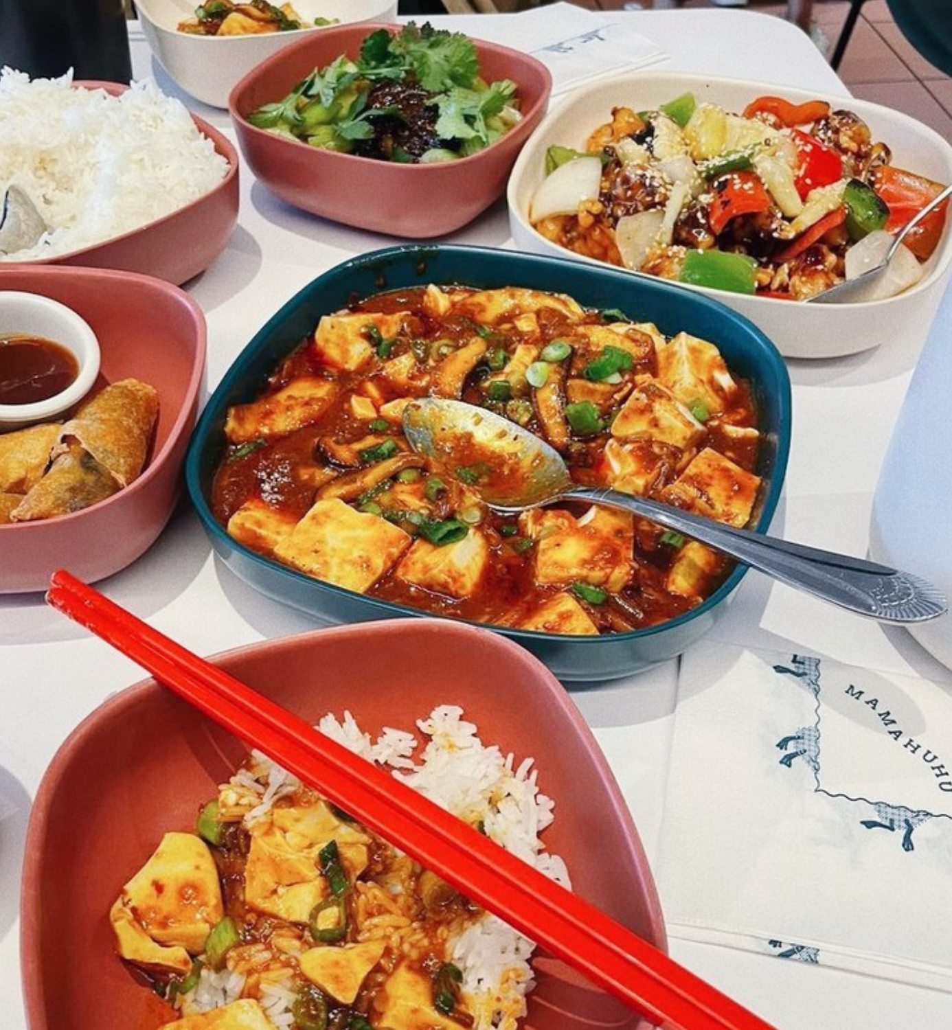 A photo of a meal at Mamahuhu, featuring Mapo Tofu House Special, Sweet & Sour Chicken House Special, Cucumber Salad, Rice, and Egg Rolls. One plate has a bowl of rice topped with Mapo Tofu.