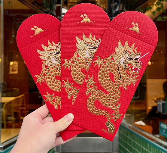 A photo of Mamahuhu's Lunar New Year 2024 Envelopes, which feature a gold dragon with sungalsses and the Mamahuhu duck logo embossed in gold on a red background. The envelopes are being held against the front of Mamahuhu's Noe Valley restaurant.