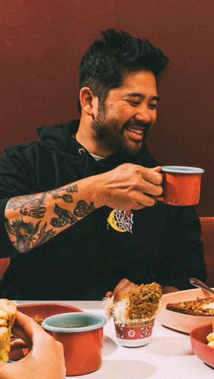 An image of a smiling Chef Brandon Jew, the co-founder of Mamahuhu. Chef Brandon is clinking his tea cup with someone out of frame and they are eating breakfast.